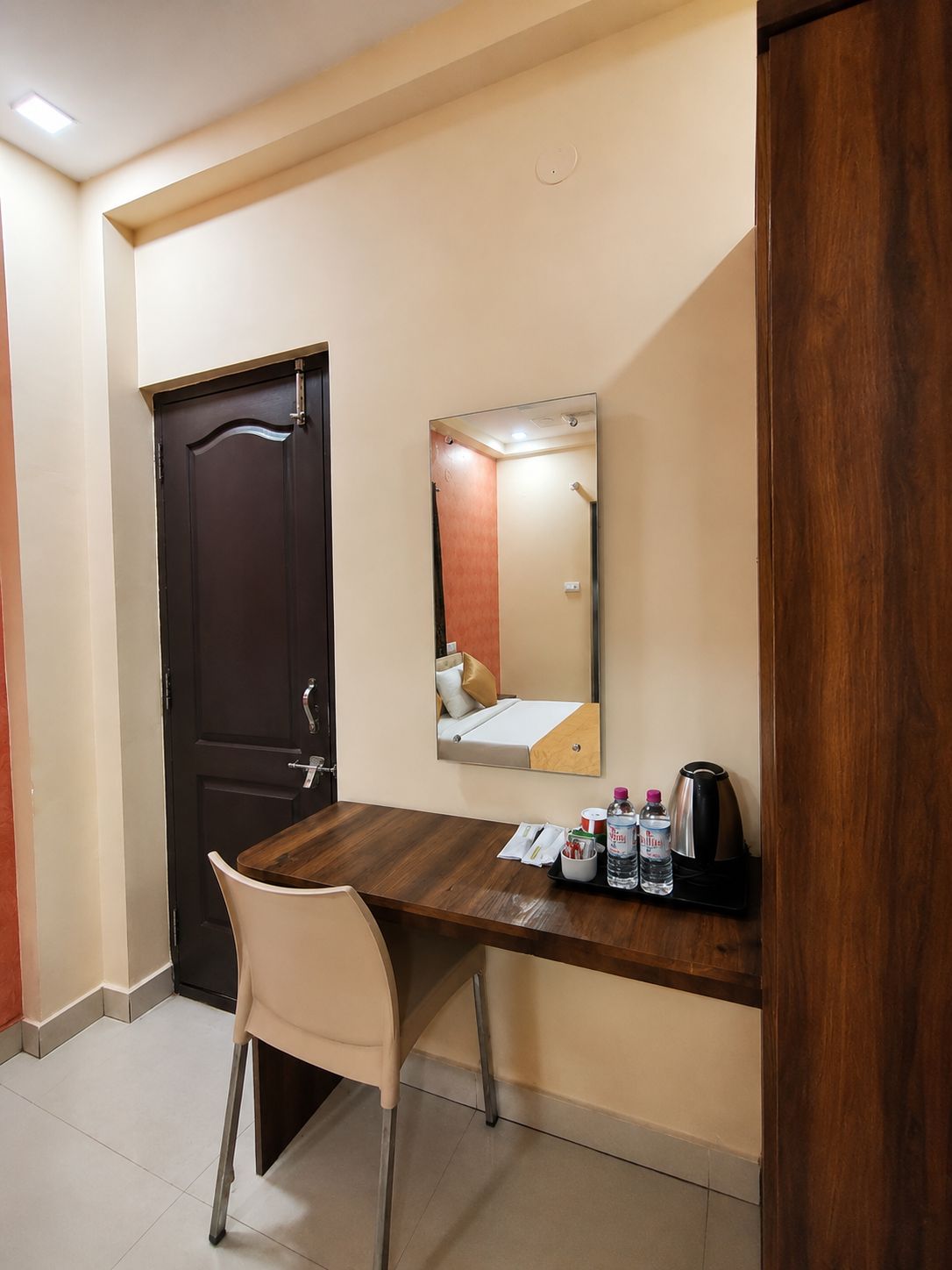 Une chambre d’hôtel avec un bureau en bois, une chaise, un lit avec une tête de lit, un miroir, une machine à café et une bouteille d’eau.