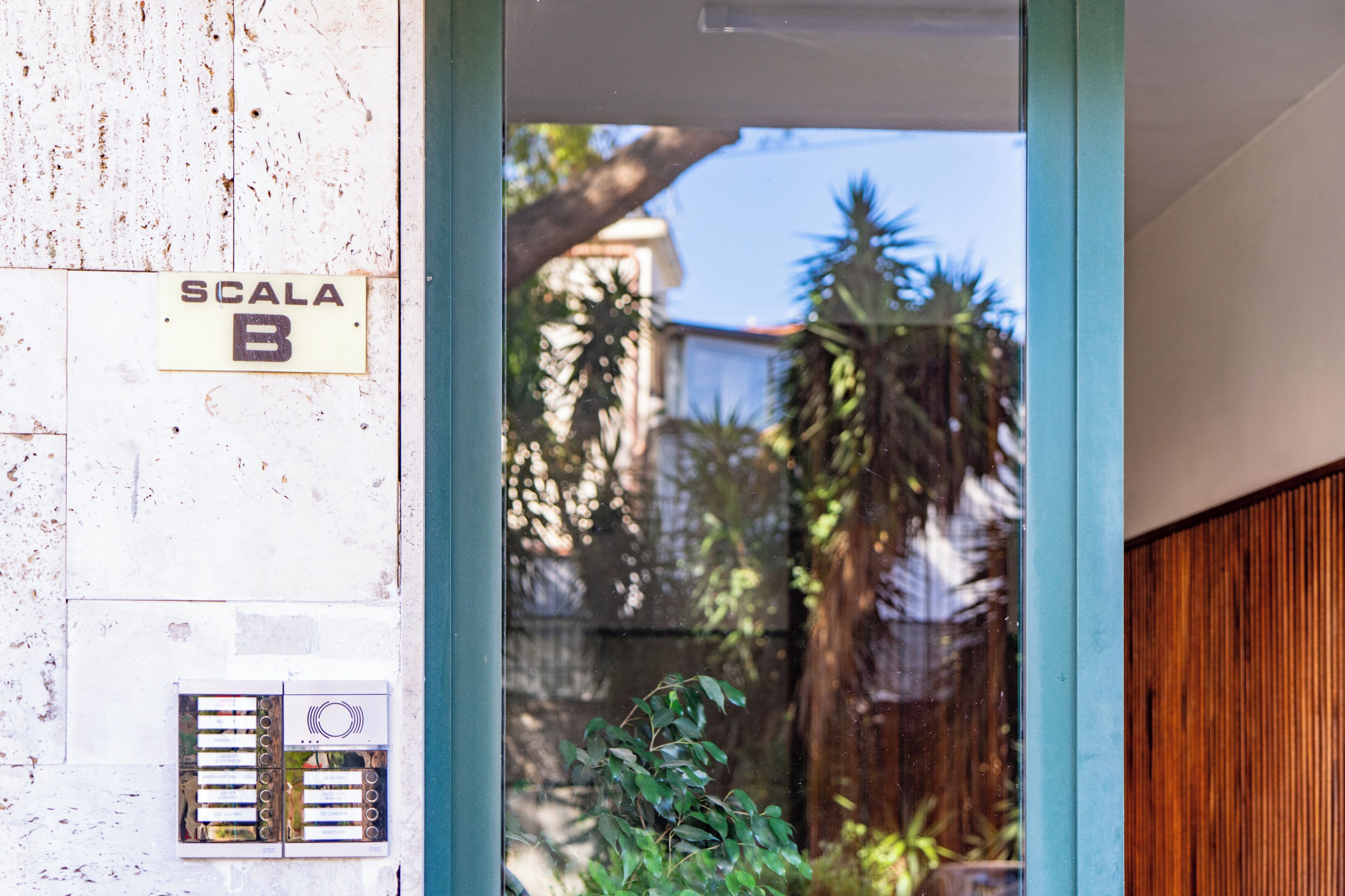 Une entrée d’immeuble avec une pancarte indiquant « SCALA B », une porte en bois et une vue sur un quartier résidentiel avec des arbres et une clôture.