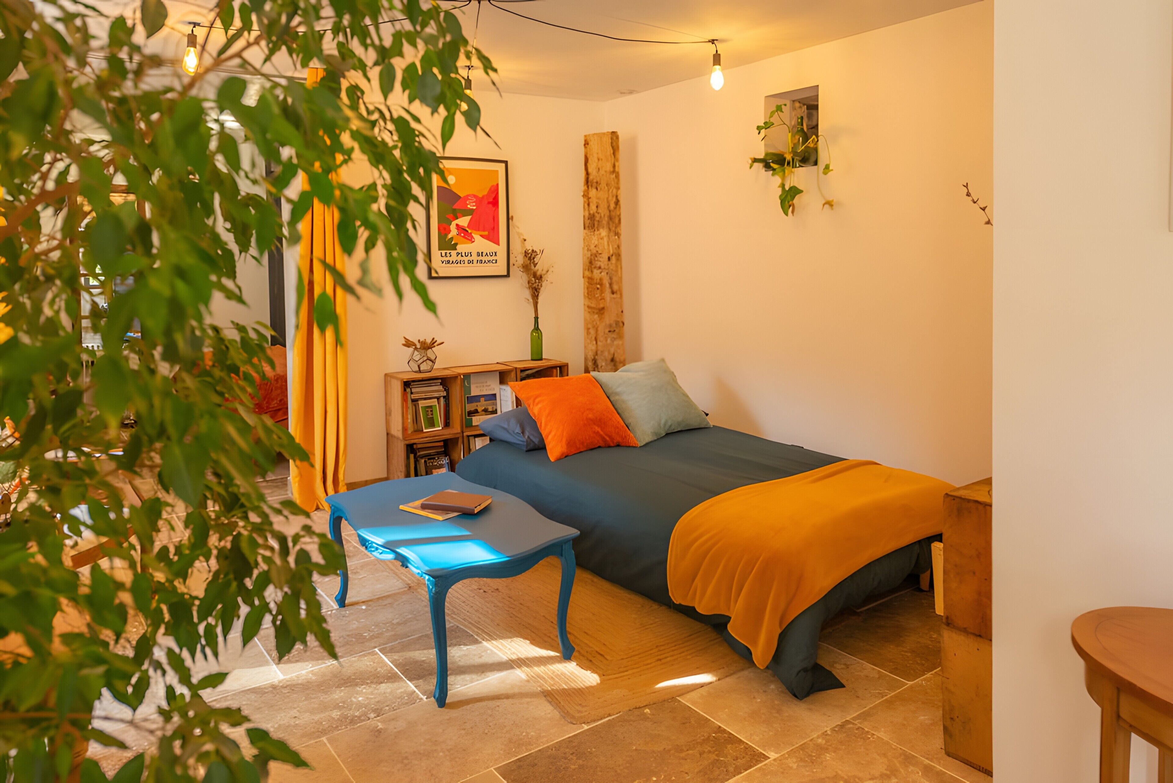 A bedroom with a bed, a blue table, a wooden side table, a plant, and a poster on the wall.