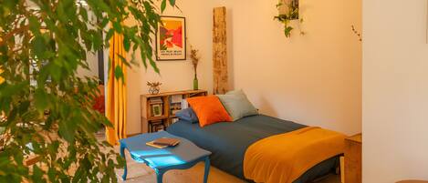 A bedroom with a bed, a blue table, a wooden side table, a plant, and a poster on the wall.