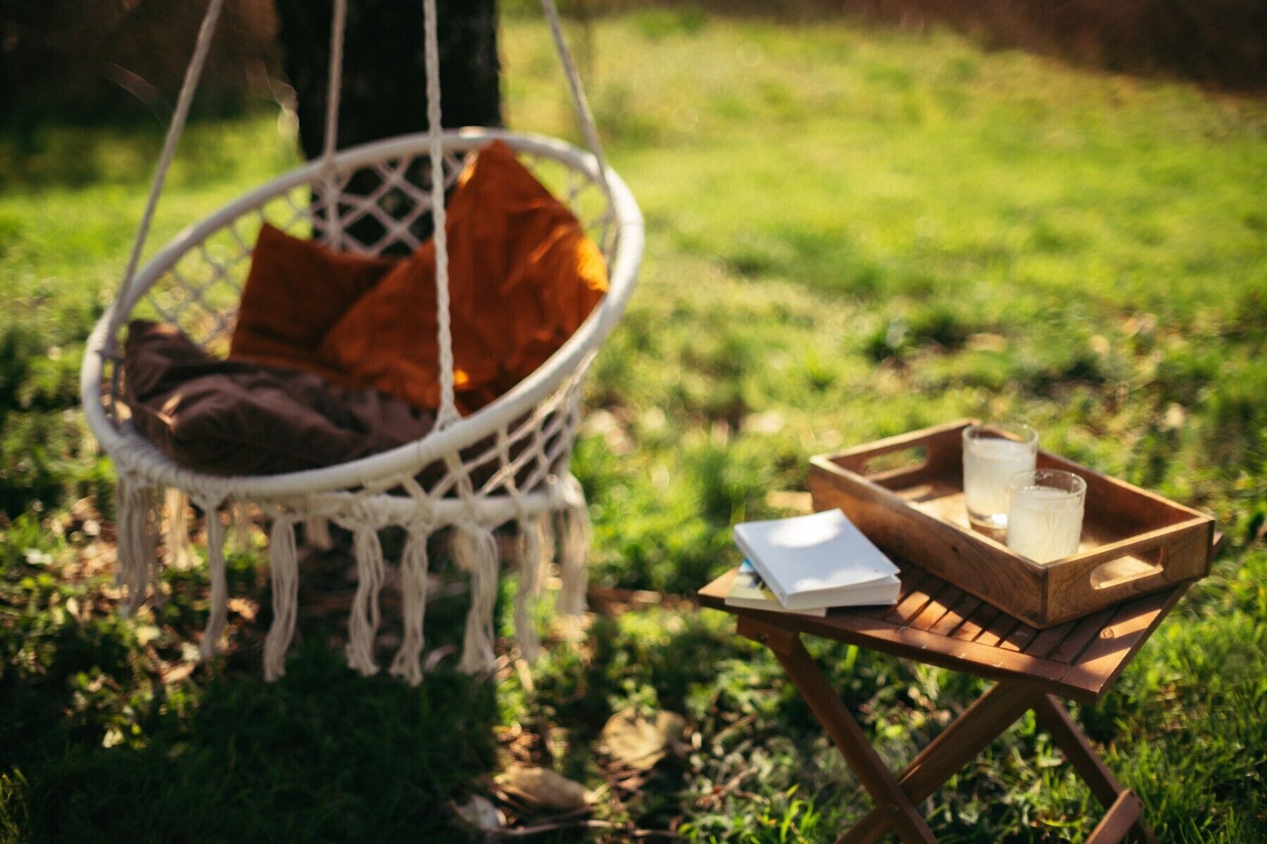 Eine Hängesessel-Stuhlsitzgelegenheit mit Kissen, ein Holztablett mit Buch und Brille sowie ein kleiner Tisch in einer grünen Umgebung im Freien.