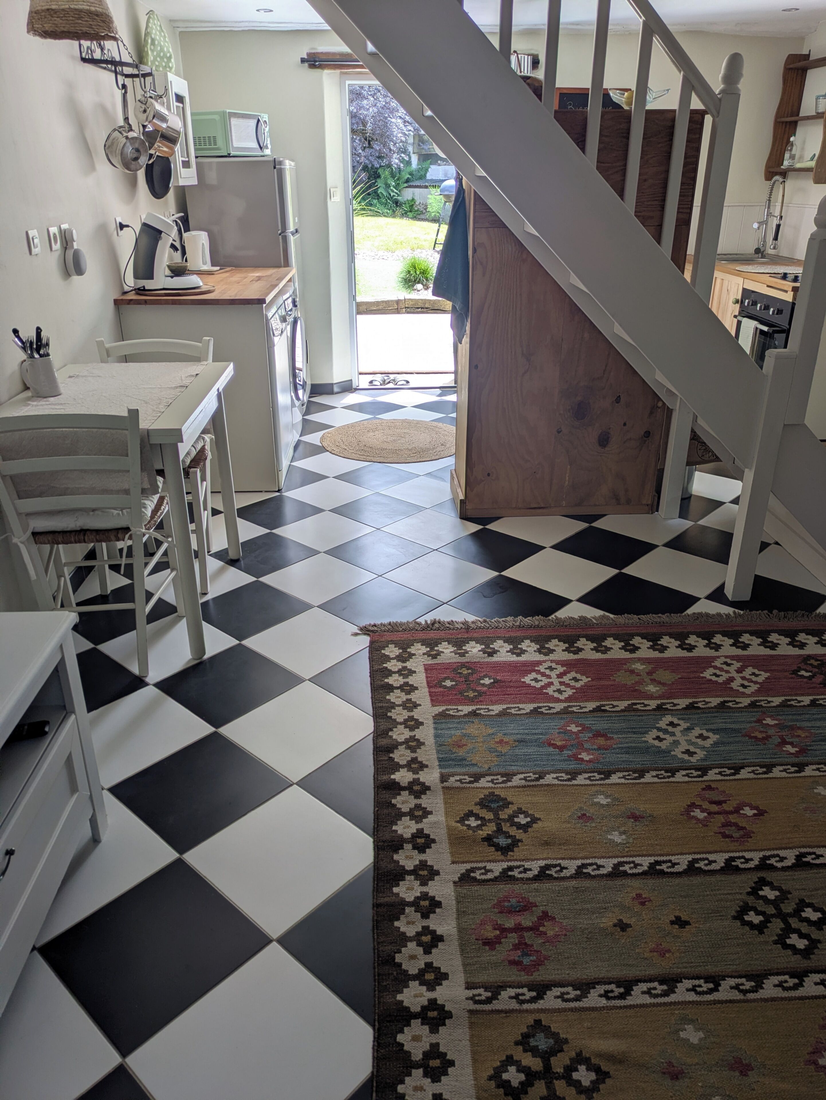 Een keuken met een geruite vloer, een eettafel, stoelen, een vloerkleed en een trap naar een andere kamer.