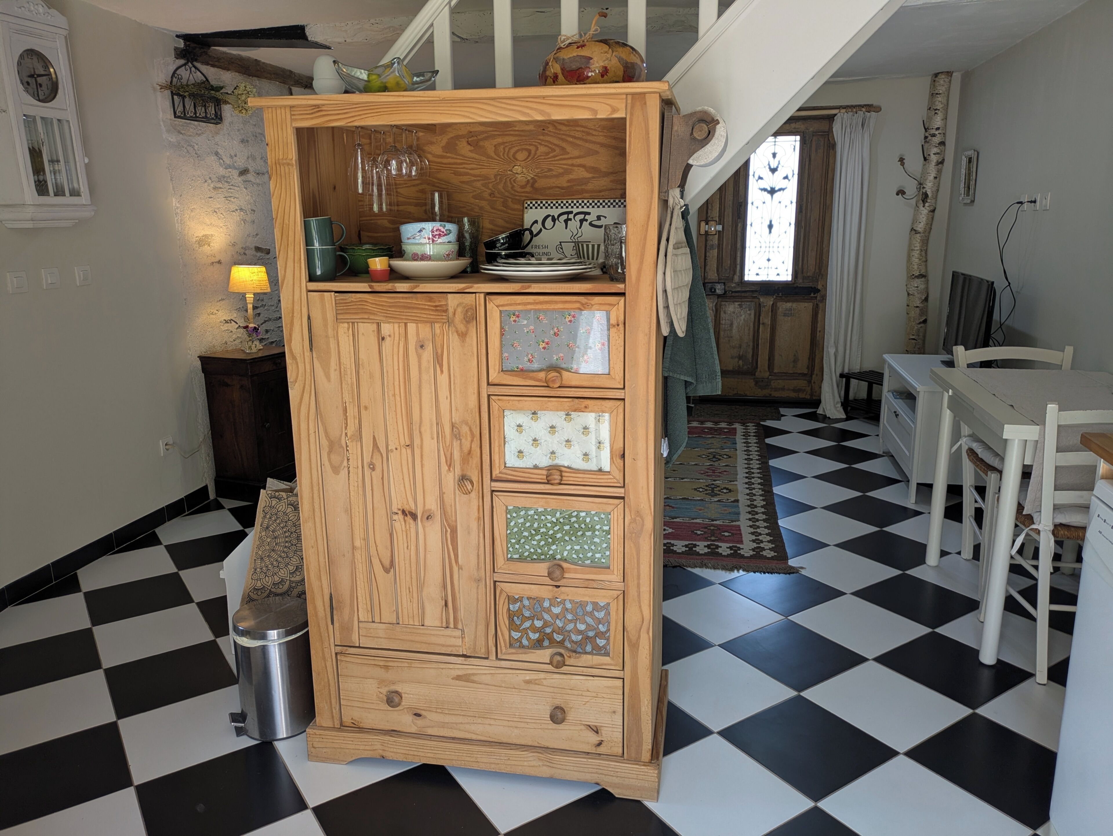 Een keuken met een geruit vloertje, een houten kast met lades, een hanglamp en een klok aan de muur.