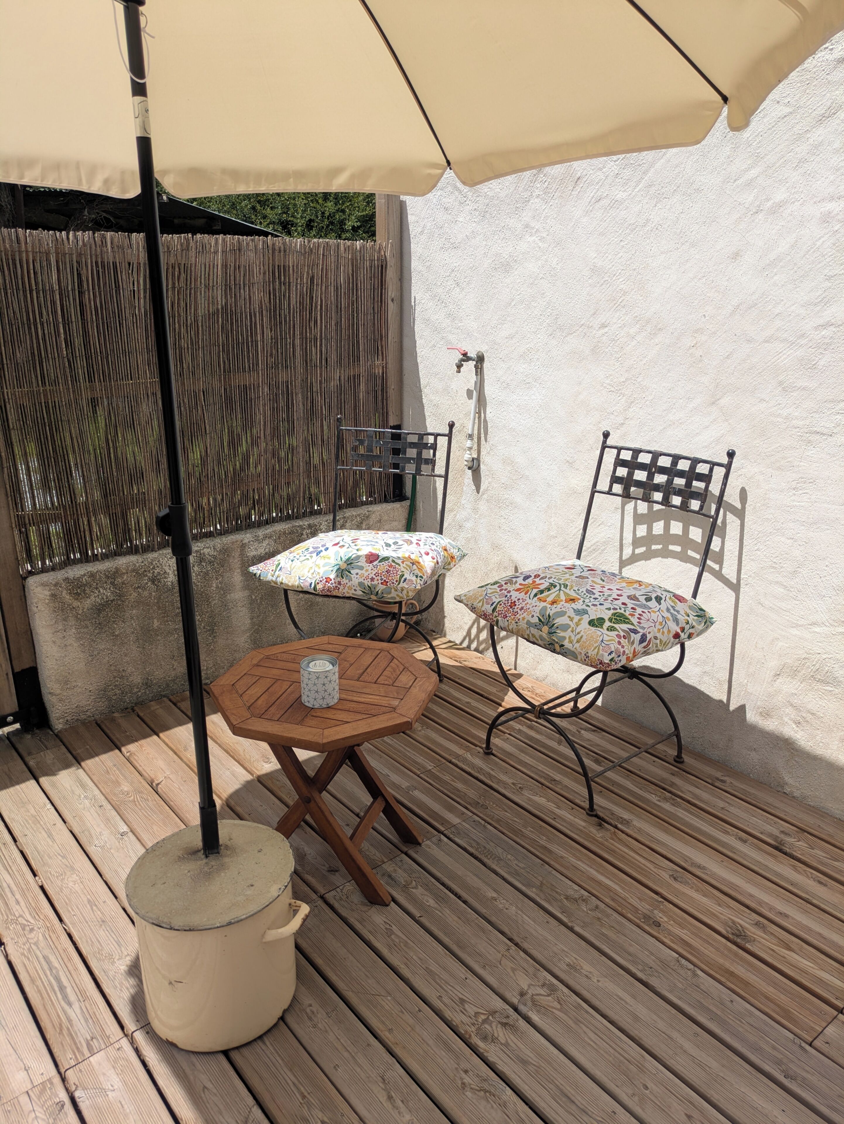 An outdoor patio with a round wooden table, two chairs with floral cushions, a beige umbrella, and a metal chair against a white wall.