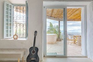 A room with a guitar, a bench, a window with metal bars, and a sliding glass door leading to an outdoor patio with a table and chairs. - Heated Plunge Pool | Mountain View’s (Málaga)