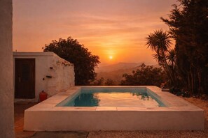 Ein rechteckiger Pool mit Blick auf den Sonnenuntergang, umgeben von Vegetation und einem Gebäude mit einer Tür und einem roten Gegenstand.