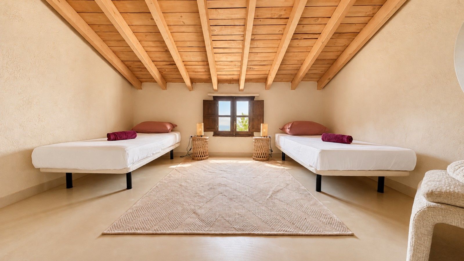 A bedroom with two beds, a large rug, wooden beams on the ceiling, and a window.
