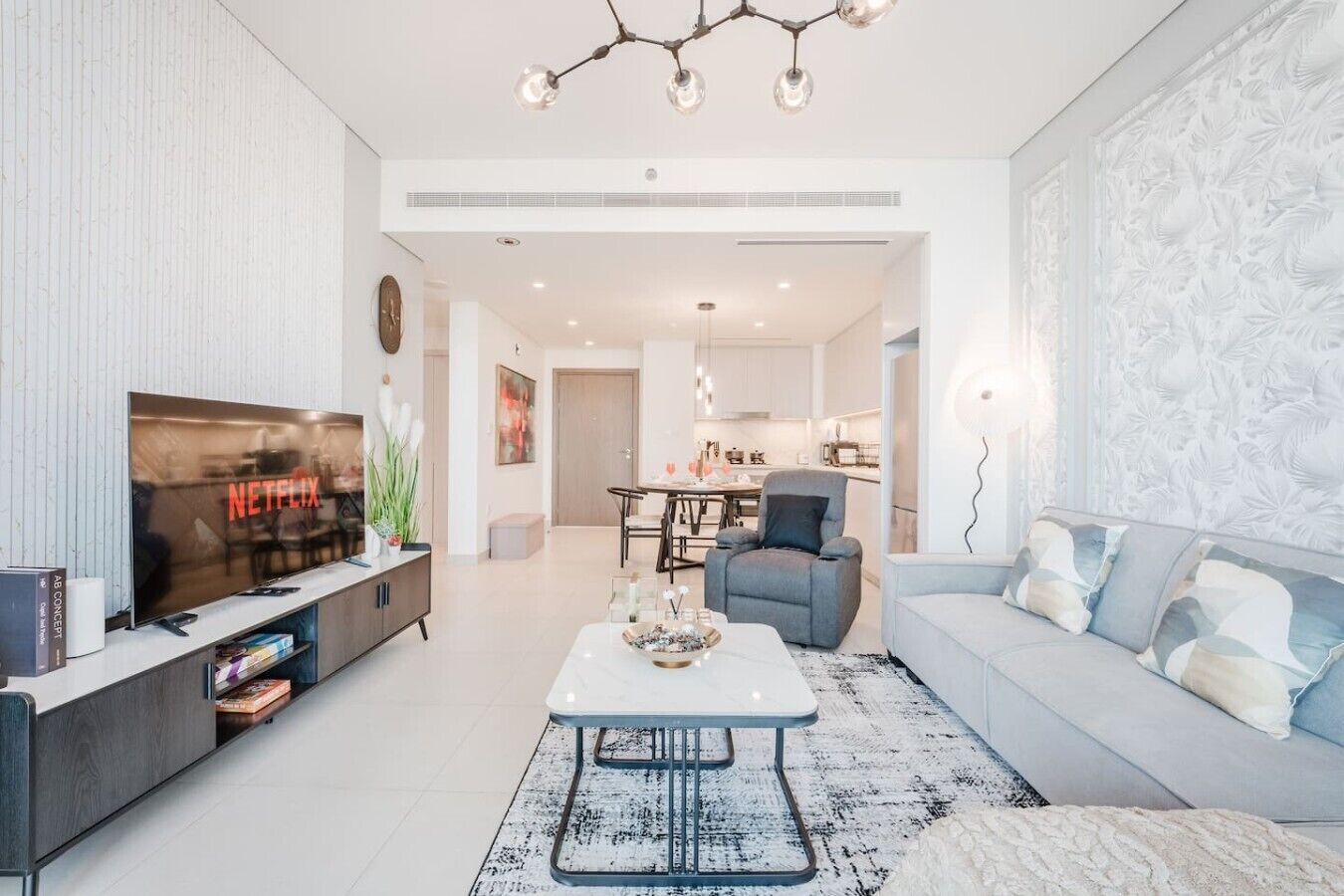 A modern living room with a flat-screen TV, a sofa, a coffee table, and a dining area in the background.