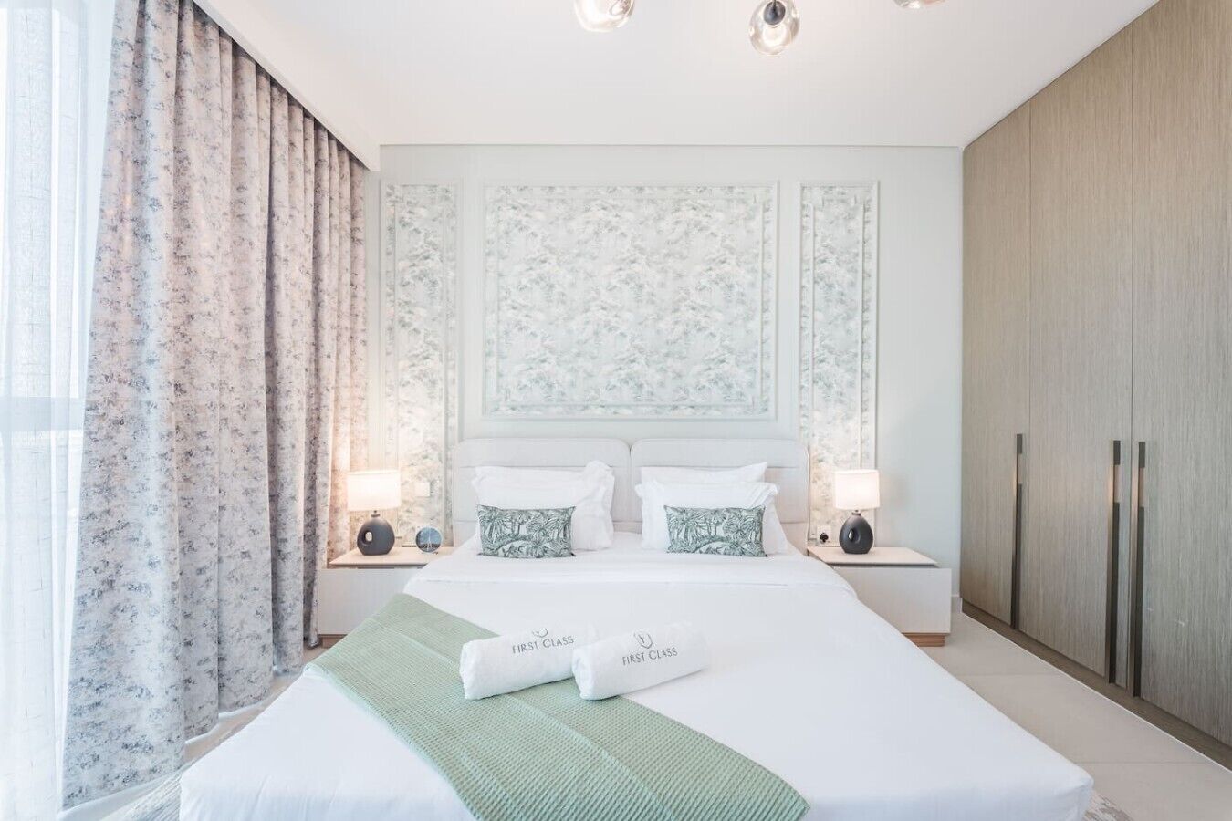 A hotel room with a large bed, two bedside tables, a nightstand, a wardrobe, and a window with curtains.