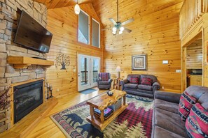 A log cabin interior with a fireplace, television, ceiling fan, and a cozy seating area. - The Toasted Turkey (Gatlinburg)