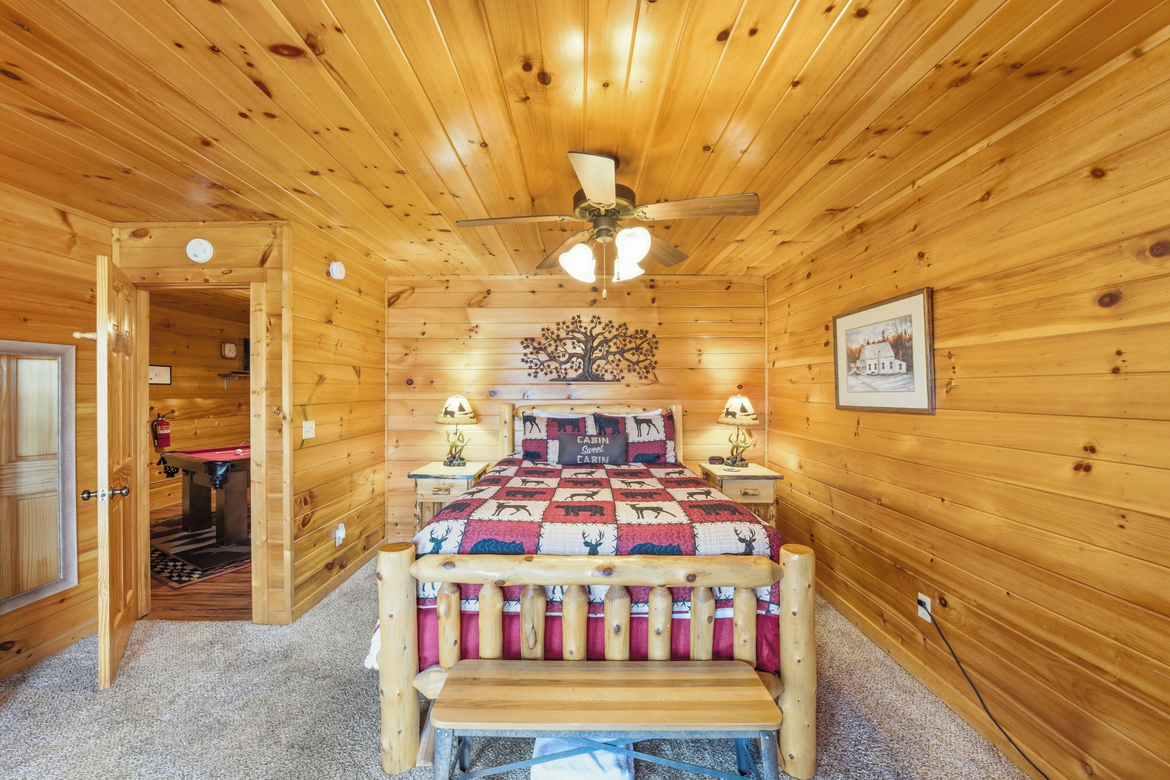 A wooden cabin bedroom with a bed, a ceiling fan, a nightstand, a lamp, and a door leading to a bathroom.