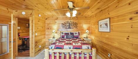 A wooden cabin bedroom with a bed, a ceiling fan, a nightstand, a lamp, and a door leading to a bathroom.