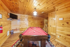 A pool table in a wooden room with a ceiling fan, a fire extinguisher, and a television mounted on the wall. - The Toasted Turkey (Gatlinburg)