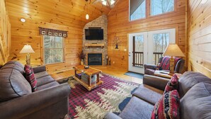 A log cabin interior with a fireplace, wooden walls, a ceiling fan, and a large window. - The Toasted Turkey (Gatlinburg)