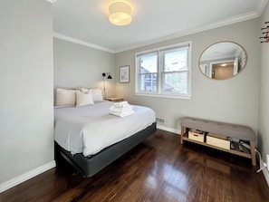 Ein Schlafzimmer mit Bett, Nachttischen, einer Bank, einem Fenster und einem an der Wand befestigten Spiegel.