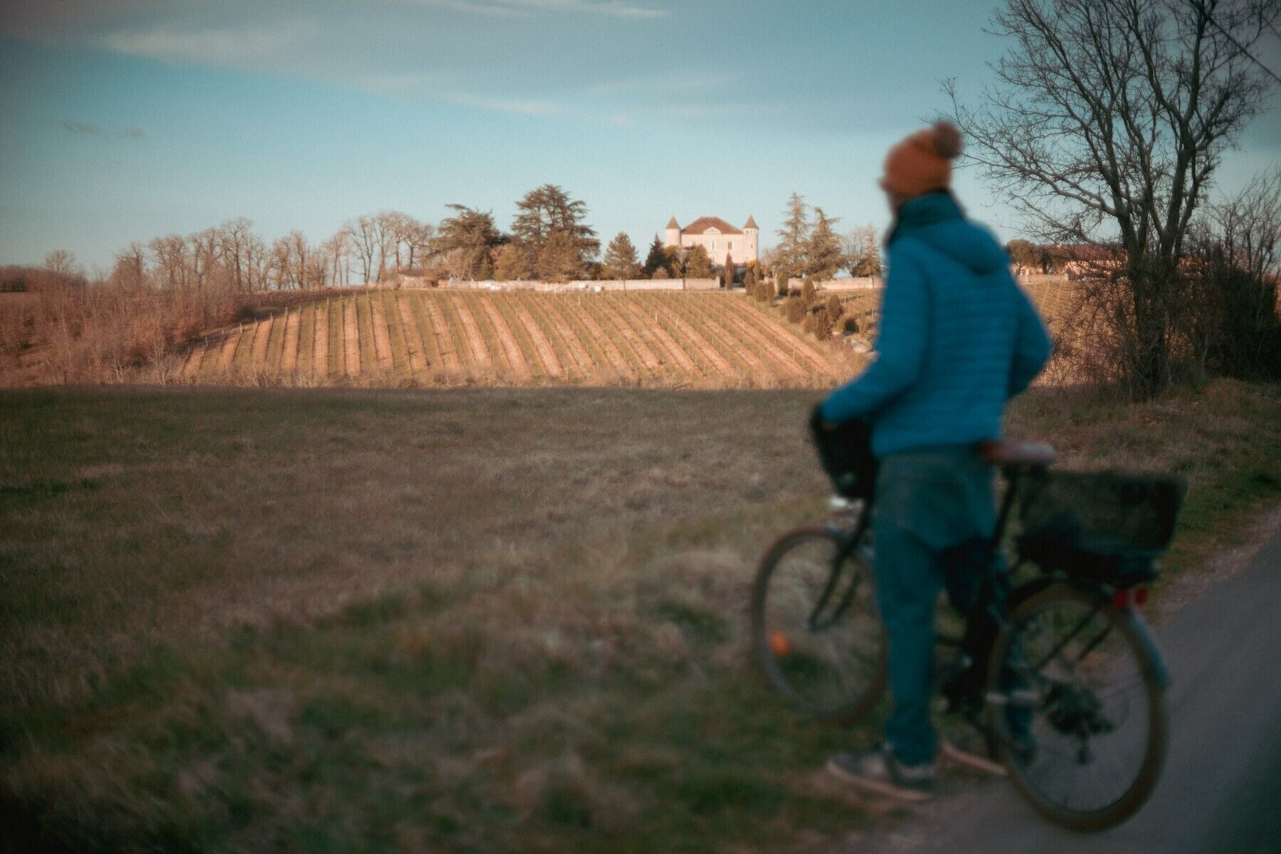 Una persona montando en bicicleta por un camino, con un campo y un edificio al fondo.