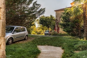 Un chemin mène à une voiture garée dans un espace verdoyant et luxuriant, avec des arbres et un bâtiment en pierre en arrière-plan.