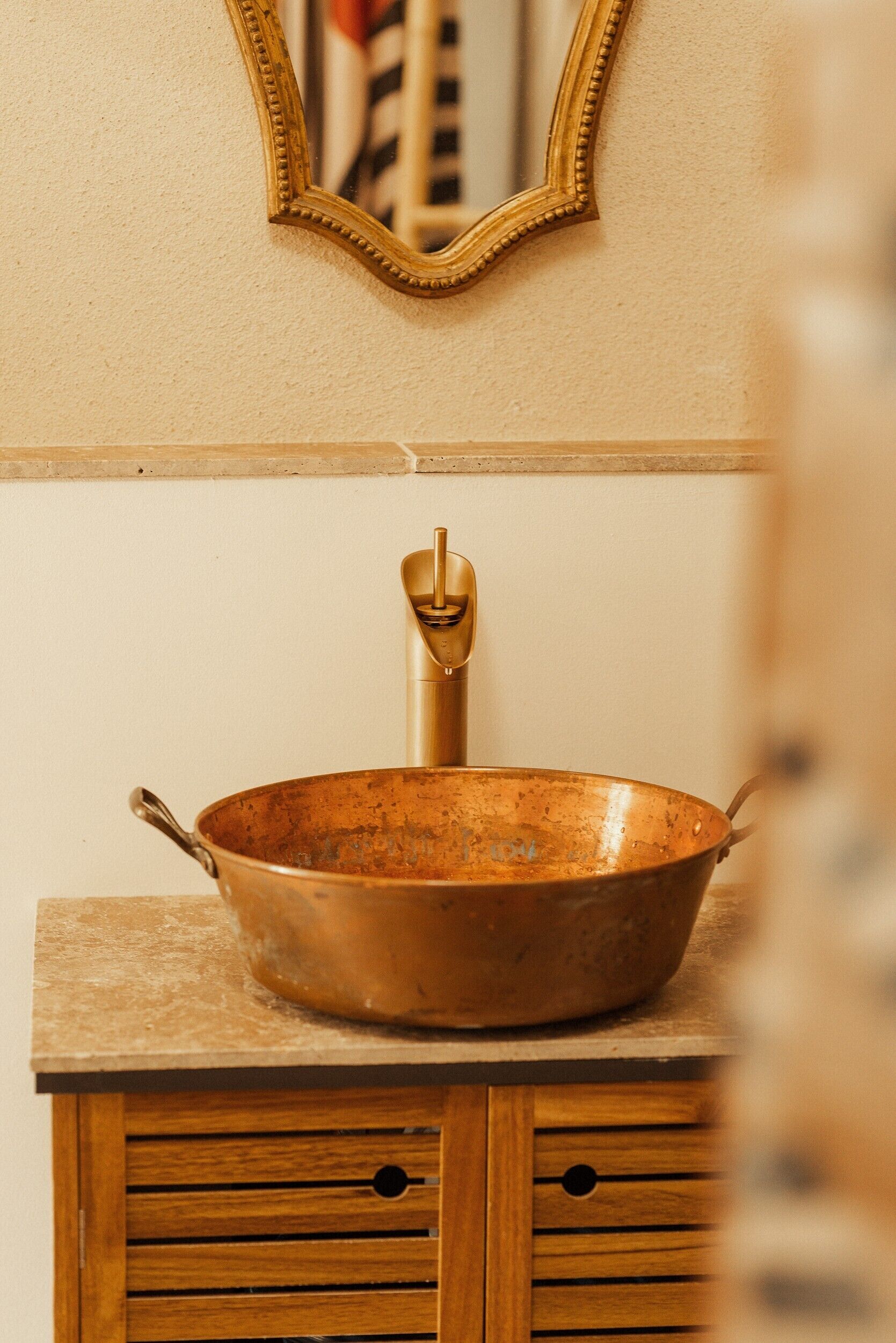 A rustic bathroom with a copper basin, wooden cabinet, and a mirror with a gold frame.