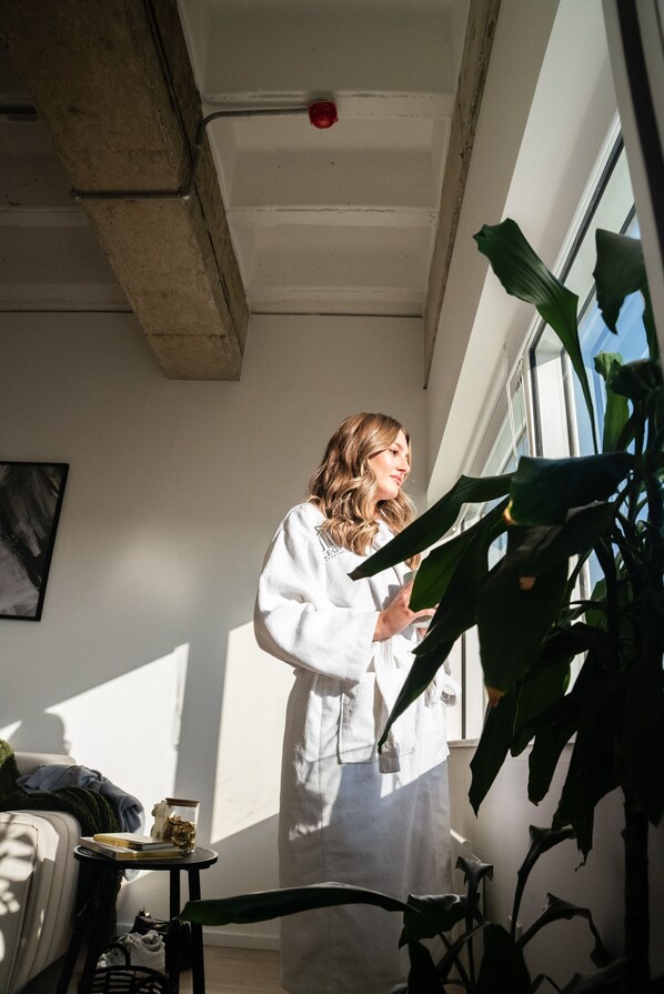 A room with a bed, a small table with a book and a container, a potted plant, and a window with a view of the sky.