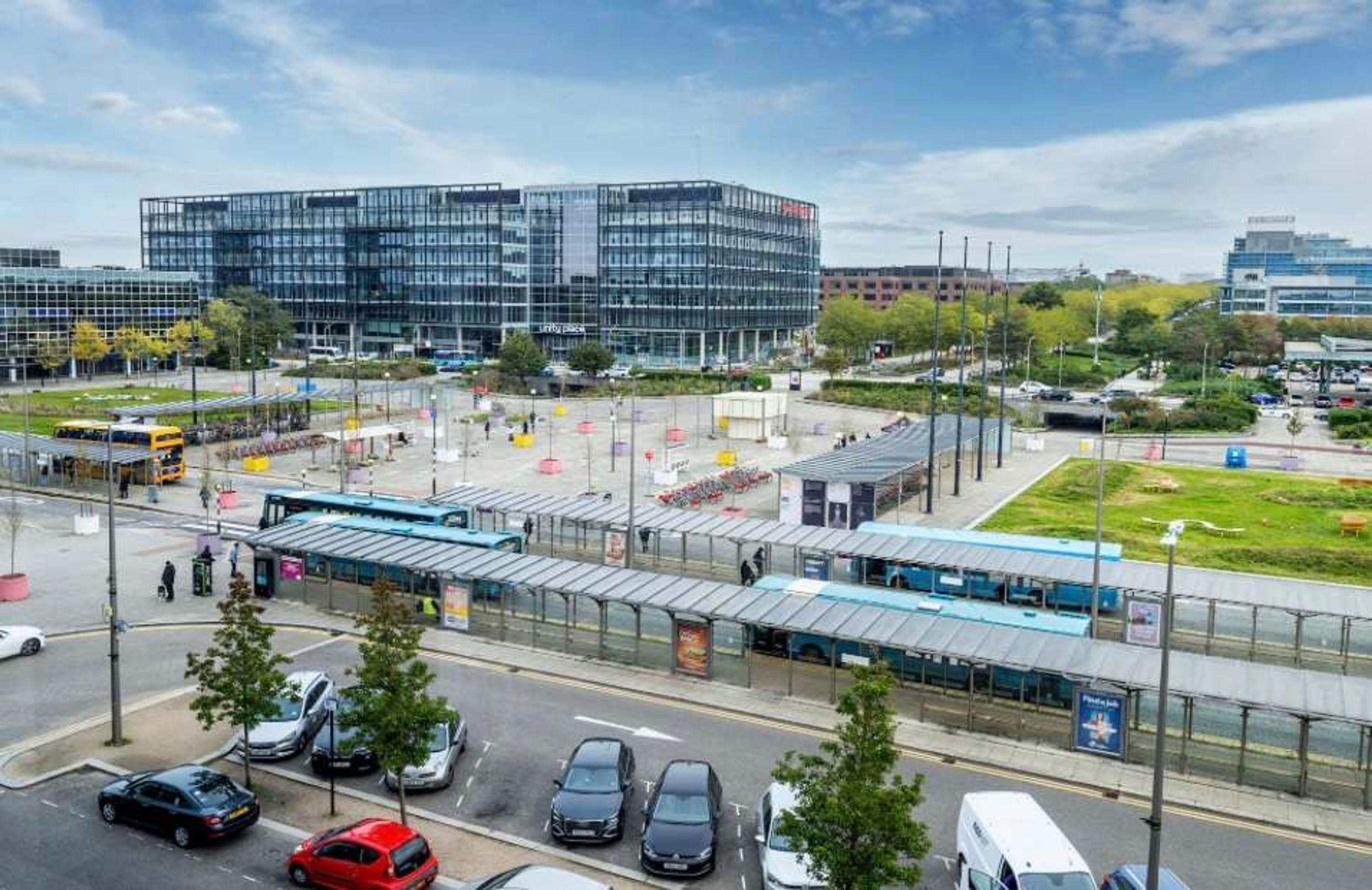 Un edificio moderno con fachada de vidrio, una estación de autobuses cubierta y un aparcamiento con coches.