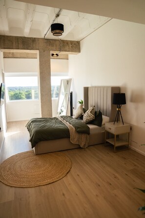 A bedroom with a bed, bedside table, lamp, and a view of greenery through a window.
