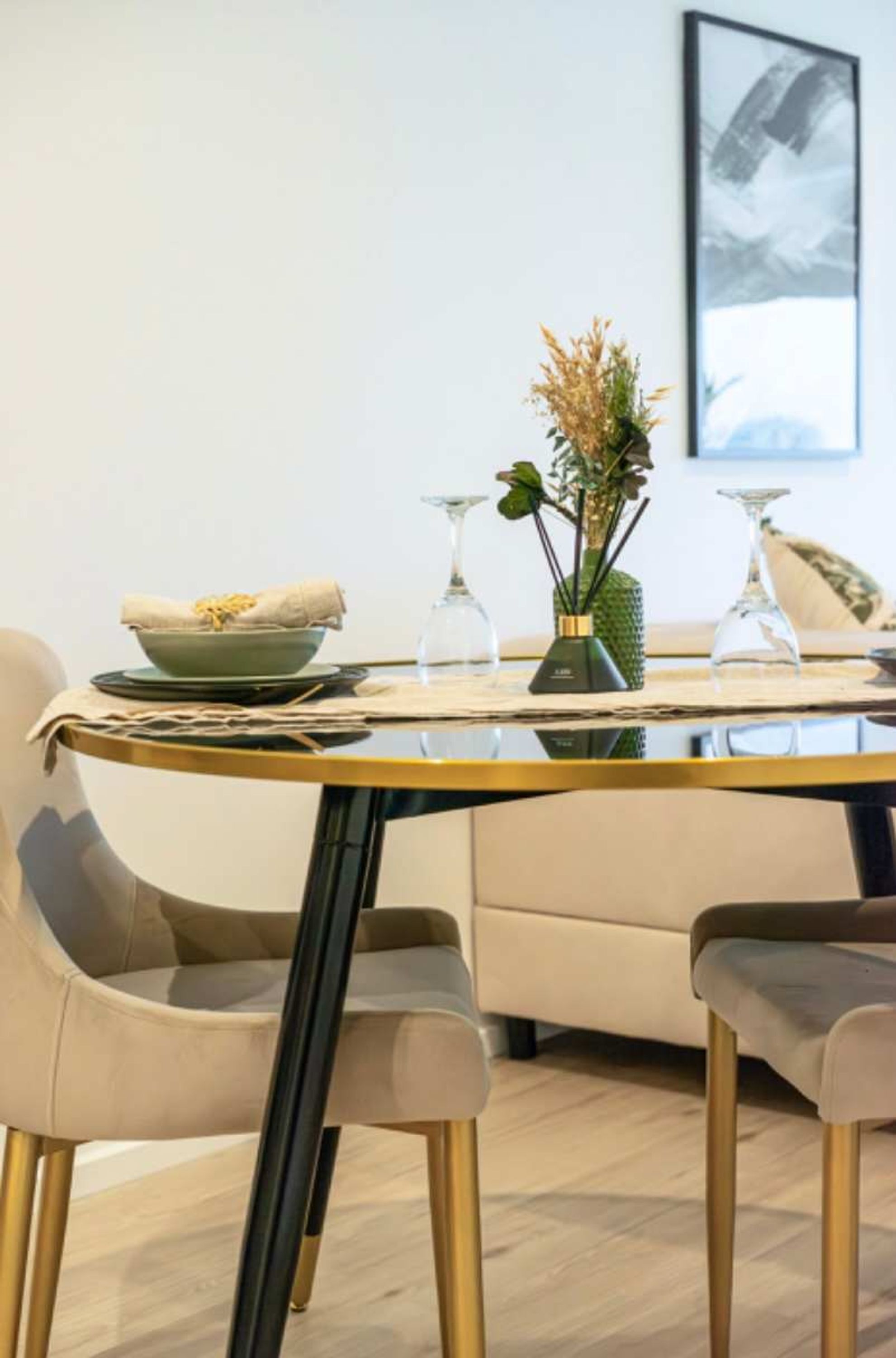 Un comedor con una mesa redonda, dos sillas, un bol, un jarrón con plantas y un cuadro en la pared.
