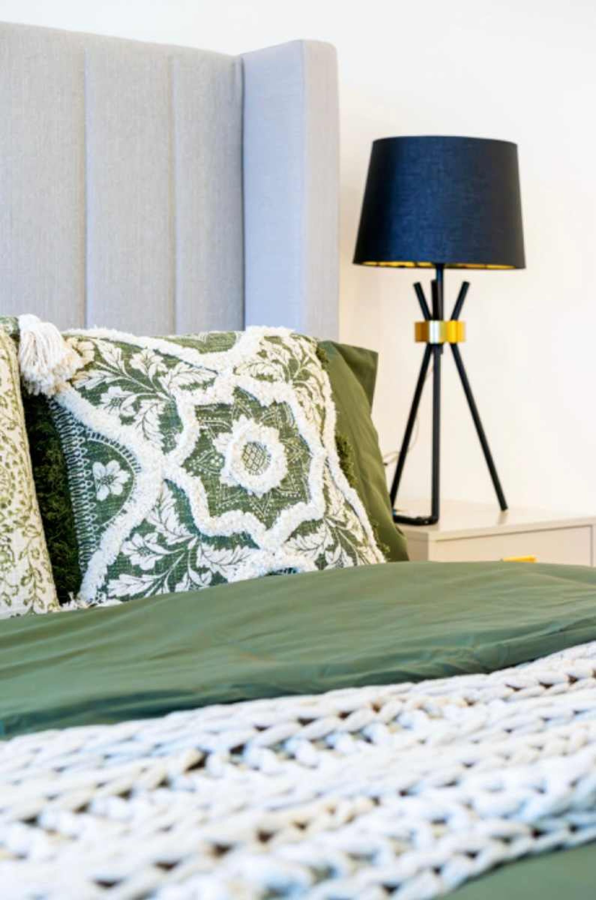 Una cama con una colcha verde, un plaid blanco de punto y almohadas estampadas. Una lámpara de pie negra con base dorada está al lado de la cama.