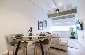 A modern living room with a glass dining table, two chairs, a sofa, and a TV mounted on the wall.