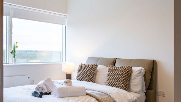 A modern bedroom with a large bed, a window with a view, a pendant light, and exposed concrete beams. - Station Elan Sleek 2-bed Apartment in Central MK (Milton Keynes)