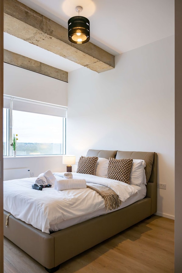 A modern bedroom with a large bed, a window with a view, a pendant light, and exposed concrete beams.