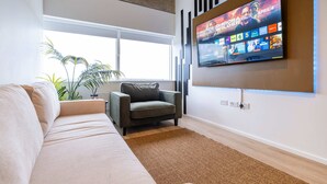 A modern living room with a sofa, a grey armchair, a flat-screen TV mounted on the wall, and a large window. - Station Elan Sleek 2-bed Apartment in Central MK (Milton Keynes)