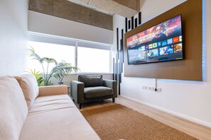 A modern living room with a sofa, a grey armchair, a flat-screen TV mounted on the wall, and a large window.