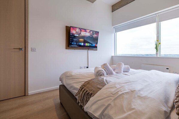 A modern hotel room with a large bed, a TV mounted on the wall, a window with a view, and a door. - Station Elan Sleek 2-bed Apartment in Central MK (Milton Keynes)