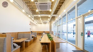 A modern interior with wooden tables and seating, large windows, and visible ceiling pipes.