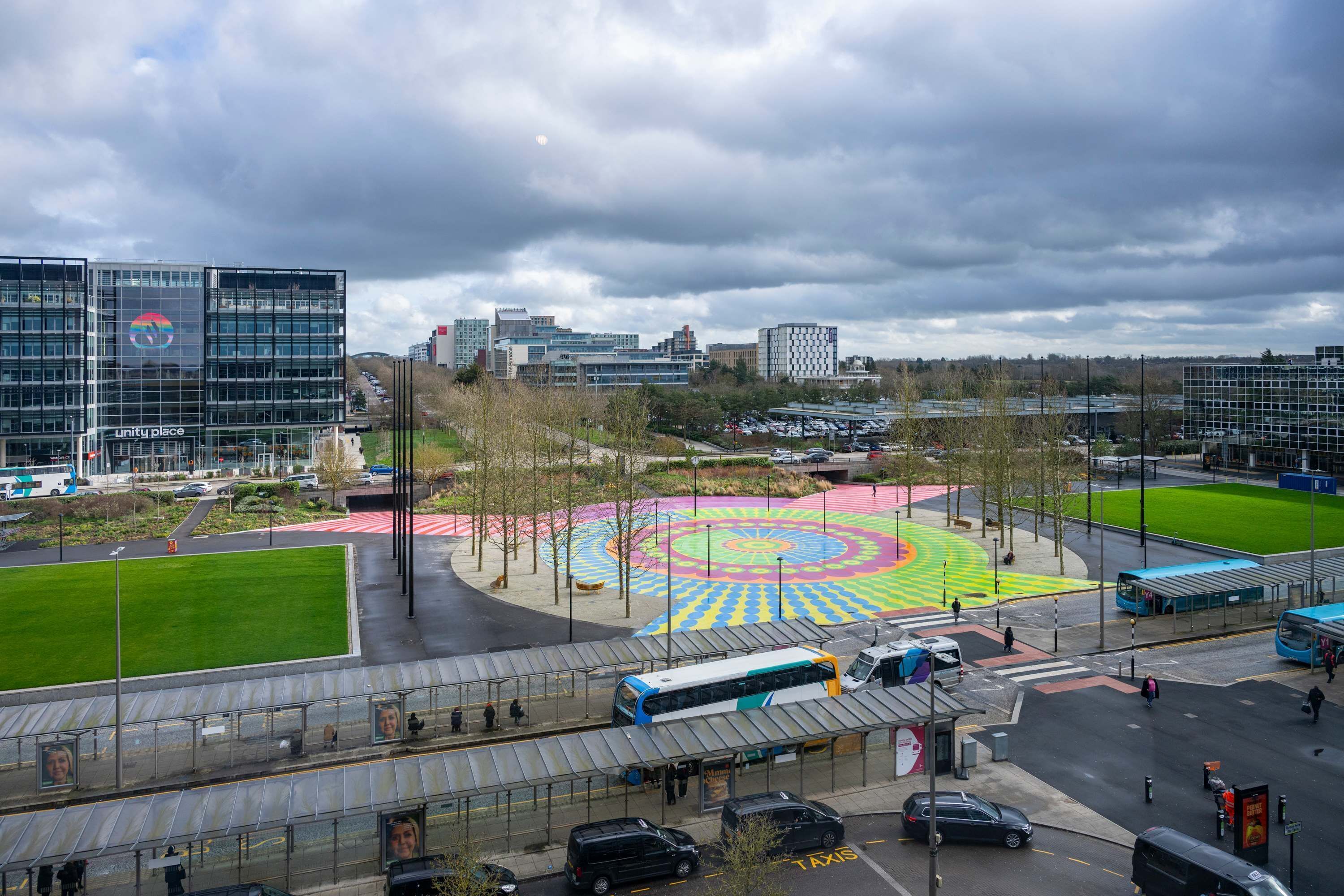 Un quartier urbain moderne comprenant une gare routière, une place circulaire aux couleurs vives et des bâtiments environnants.