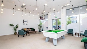 A pool table in a modern lobby with hanging lights, seating, and a view through large windows.
