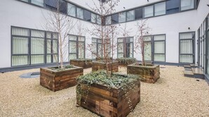 A courtyard with wooden planters, a building with large windows, and a gravel ground. - Entire Rental Unit in Linford Wood (Milton Keynes)