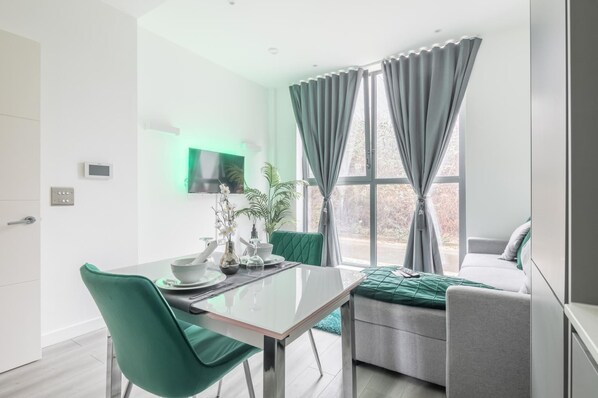 Una sala de estar moderna con una mesa de comedor de vidrio, sillas verdes, un sofá gris y un ventanal con cortinas.