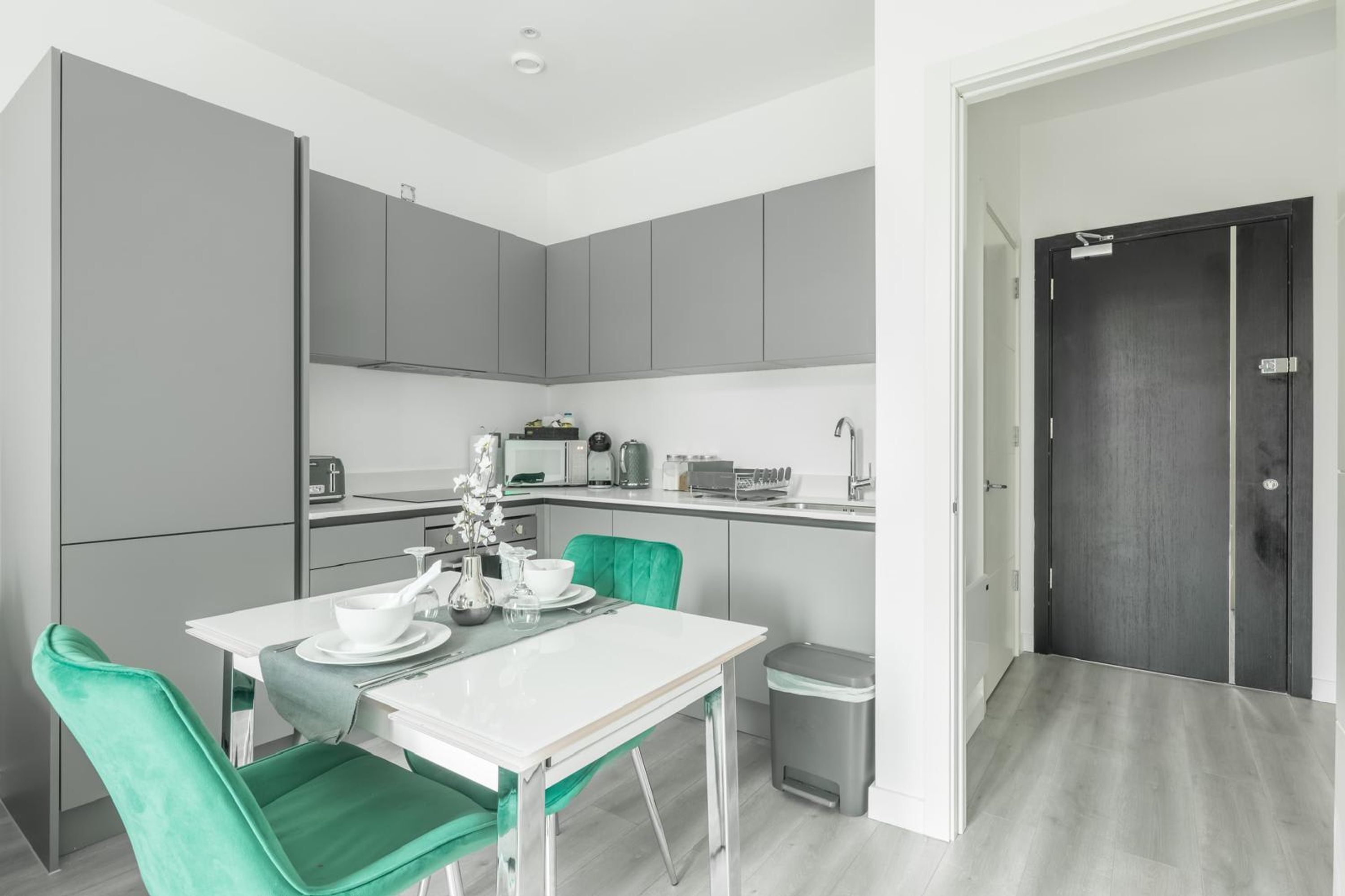 A modern kitchen with grey cabinets, a white countertop, a dining table with green chairs, and a small trash bin.