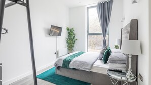 A modern hotel room with a bed, a TV, a window with curtains, a plant, and a lamp. - Entire Rental Unit in Linford Wood (Milton Keynes)