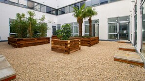 A modern building interior with a central courtyard featuring wooden planters with various plants, a pebbled floor, and large windows.