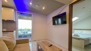 Un soggiorno moderno con un divano, un tavolino in legno, una TV montata a parete e un balcone visibile attraverso una porta di vetro.