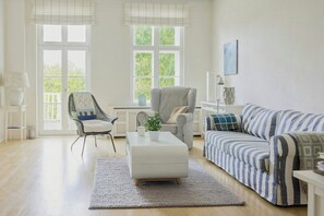 A living room with a sofa, armchair, and ottoman, a window with blinds, and a lamp. - Historic 1913 Parsonage Apartment (Butzow)