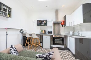 Une cuisine compacte équipée d’un four à micro-ondes, d’un grille-pain et d’un four, un petit coin salon avec un canapé et des fauteuils, et un téléviseur fixé au-dessus du comptoir.