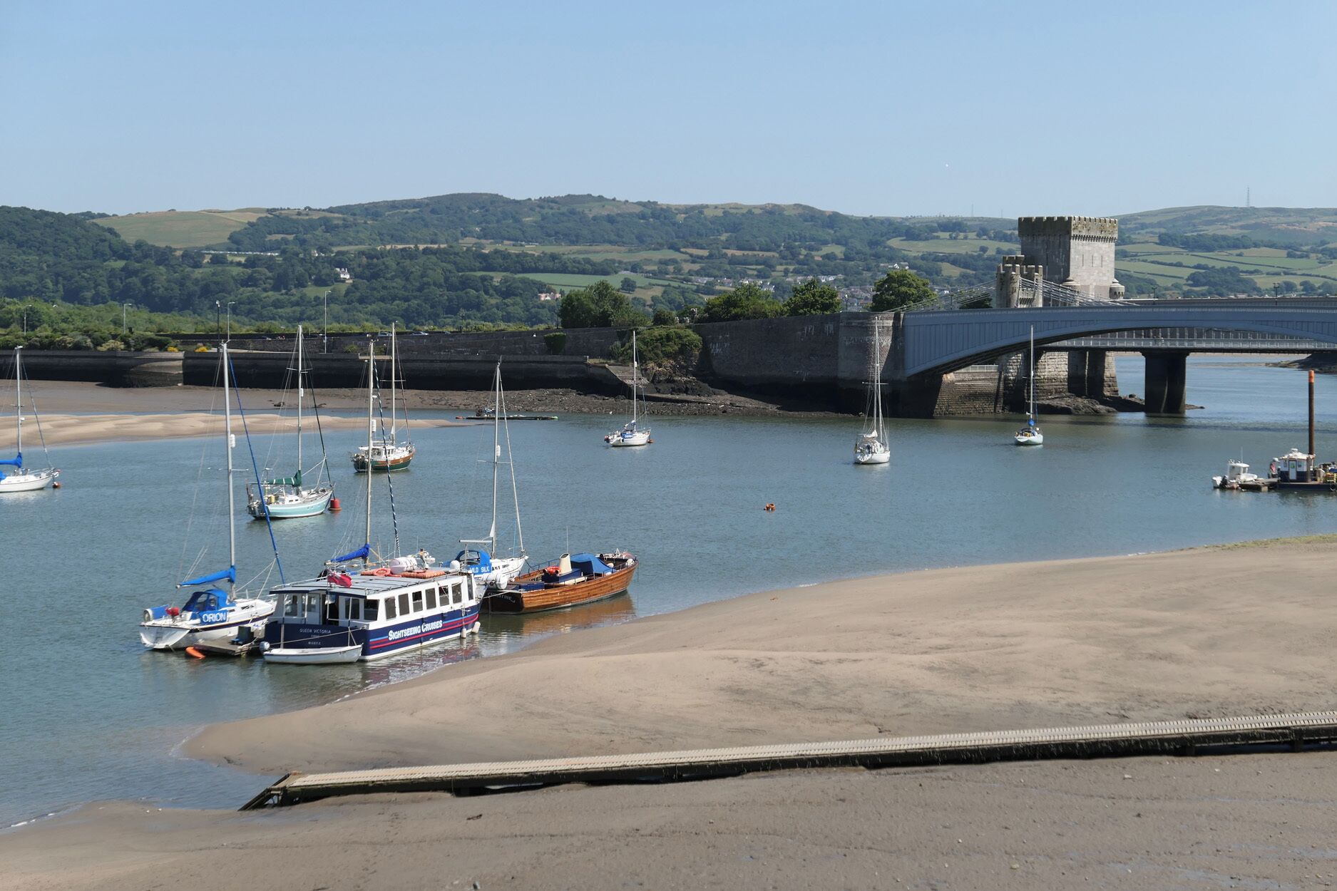 A coastal scene with boats docked near a sandy beach, a bridge, and a castle in the background.