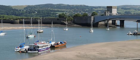 A coastal scene with boats docked near a sandy beach, a bridge, and a castle in the background.