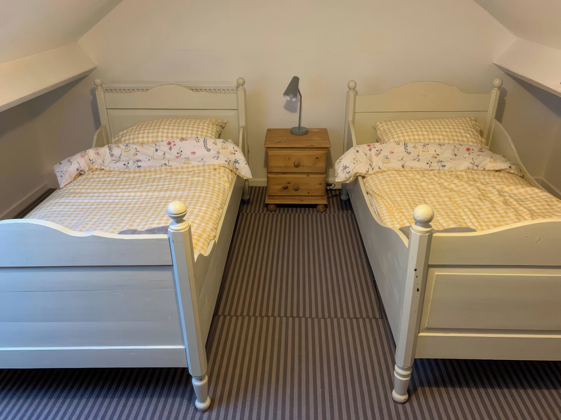 Twin beds with checkered bedding, a nightstand with a lamp, and a striped carpet.