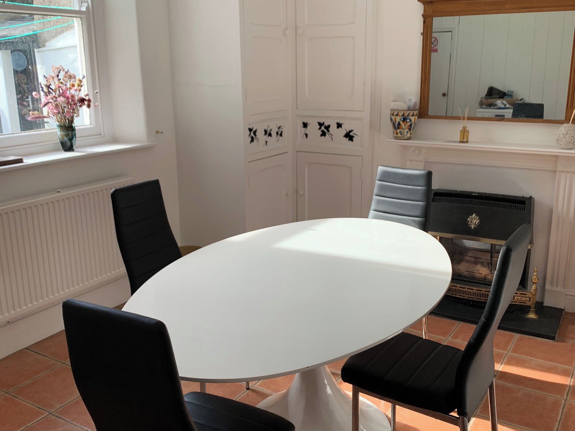 A dining area with a round white table, black chairs, a vase with flowers, a radiator, and a mirror on the wall.