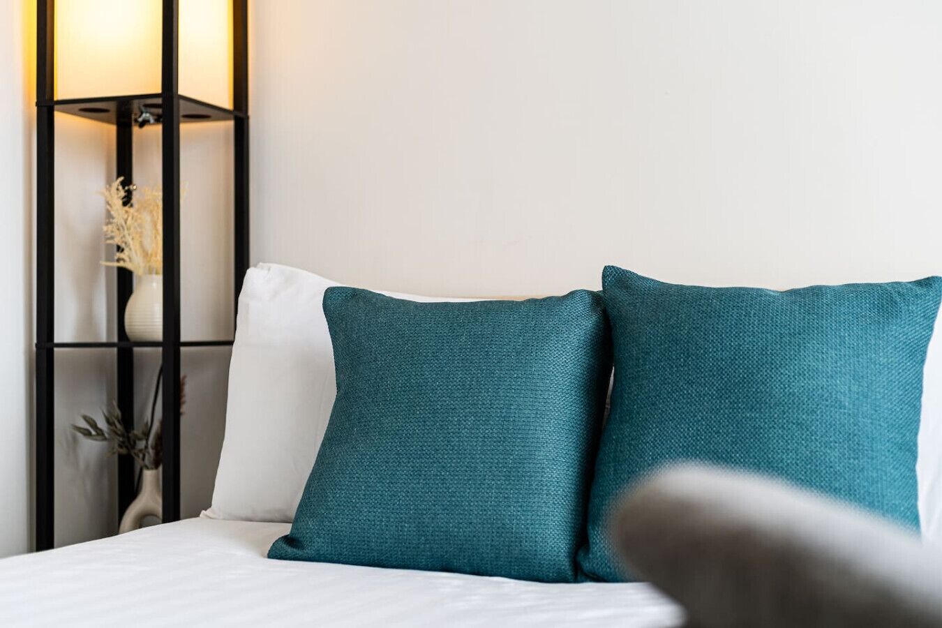 A bed with white bedding and two teal pillows, a black metal nightstand with a vase of dried flowers, and a wall-mounted lamp.