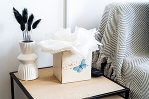 Eine weiße Vase mit schwarzen Federn, eine Taschentuchbox mit blauem Schmetterlingsmuster und ein gemustertes Stuhl.